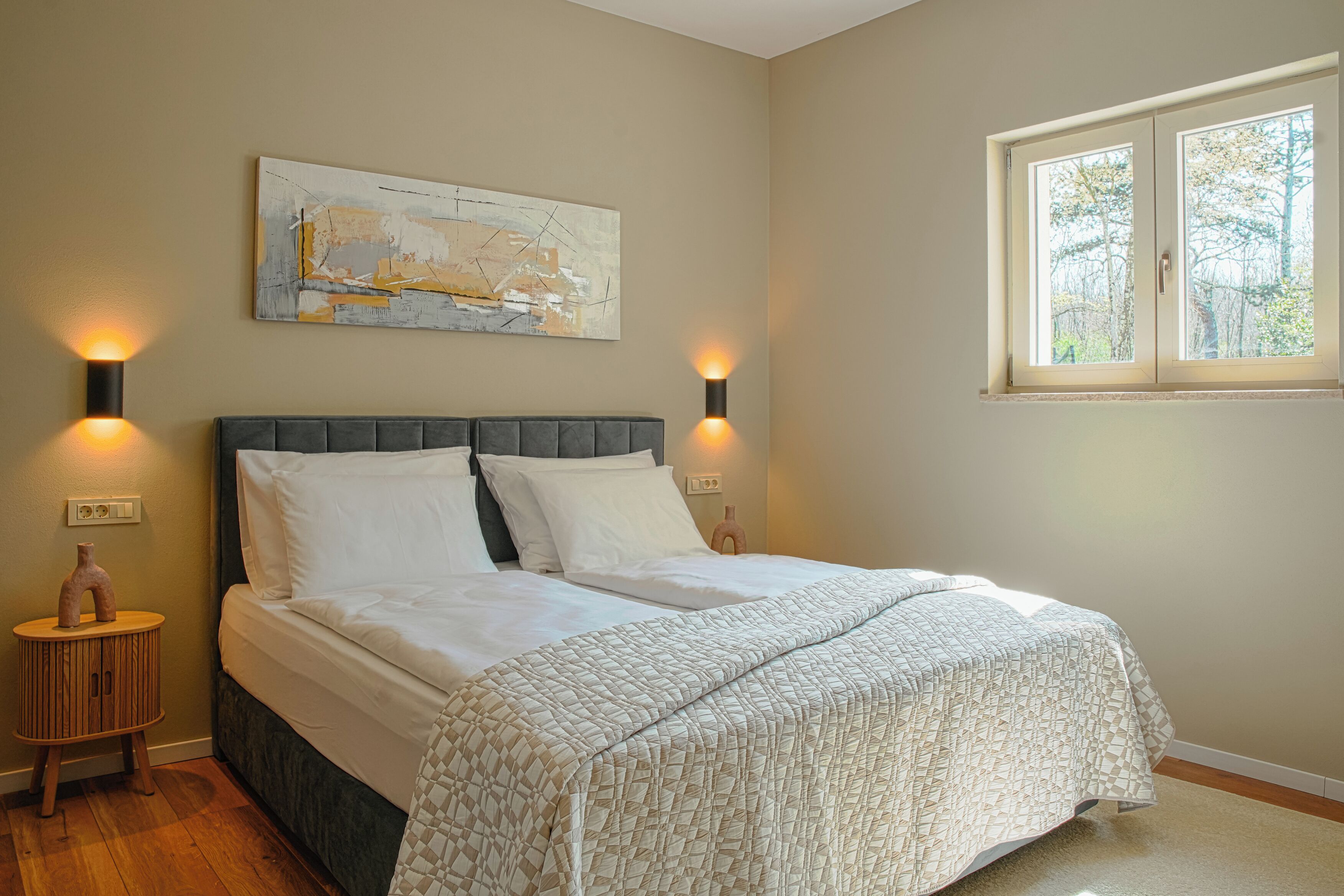 A bedroom with a bed, two bedside lamps, a wall-mounted abstract painting, and a window with a view of trees.