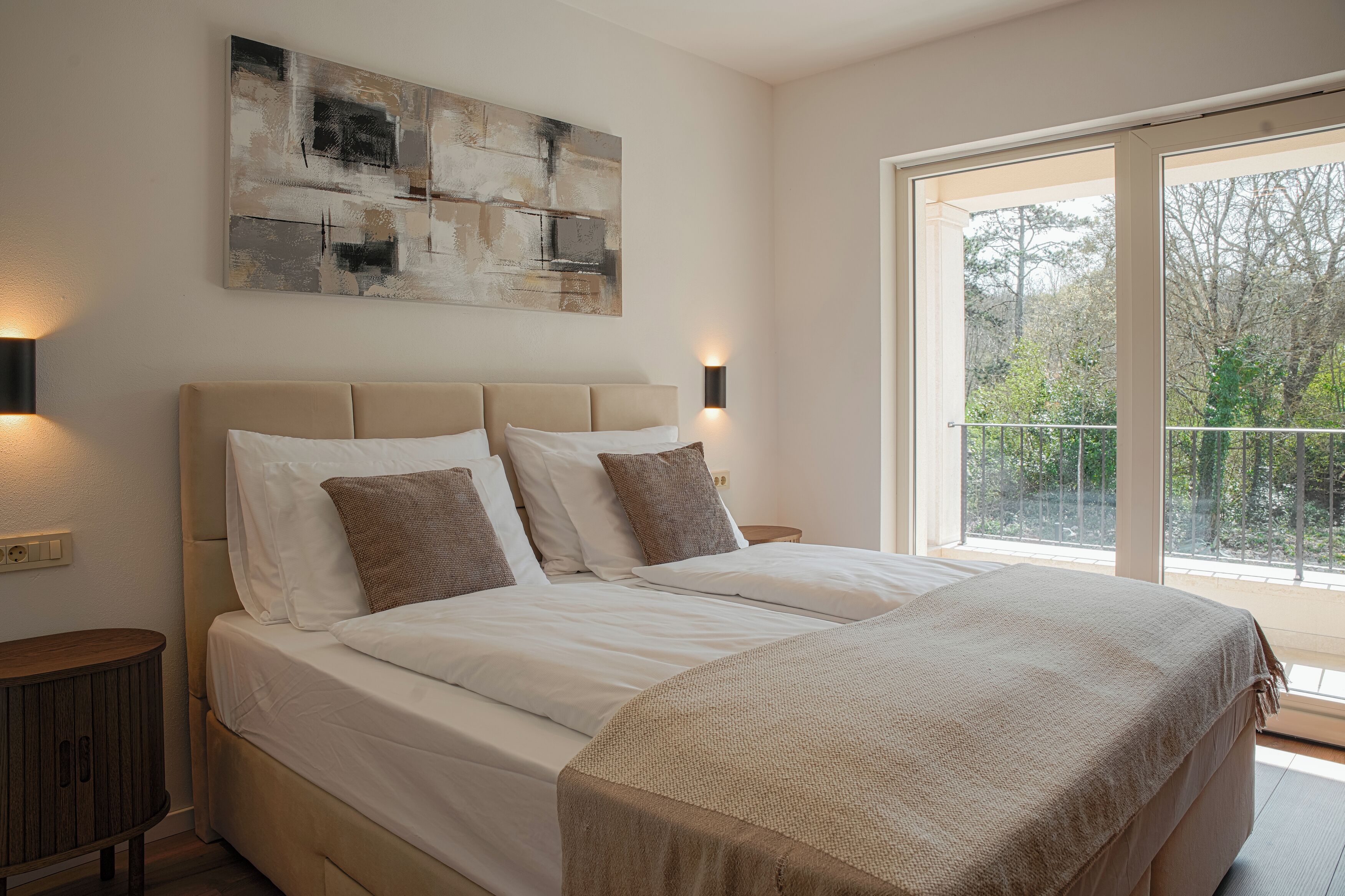 A bedroom with a large bed, a nightstand, a wall-mounted light, and a window with a view of trees.