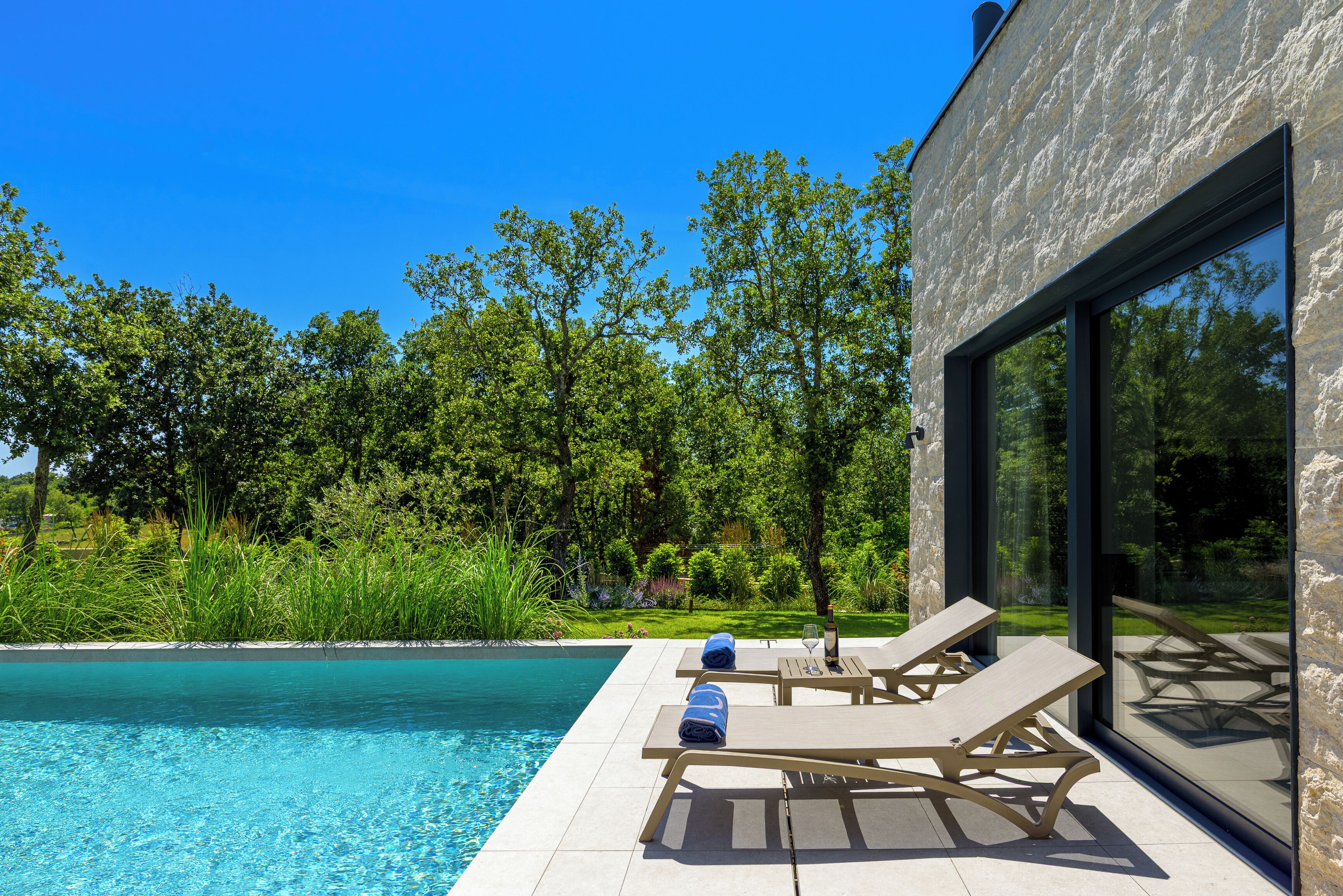 A poolside area with lounge chairs and a bottle of wine, surrounded by lush greenery and a clear blue sky.