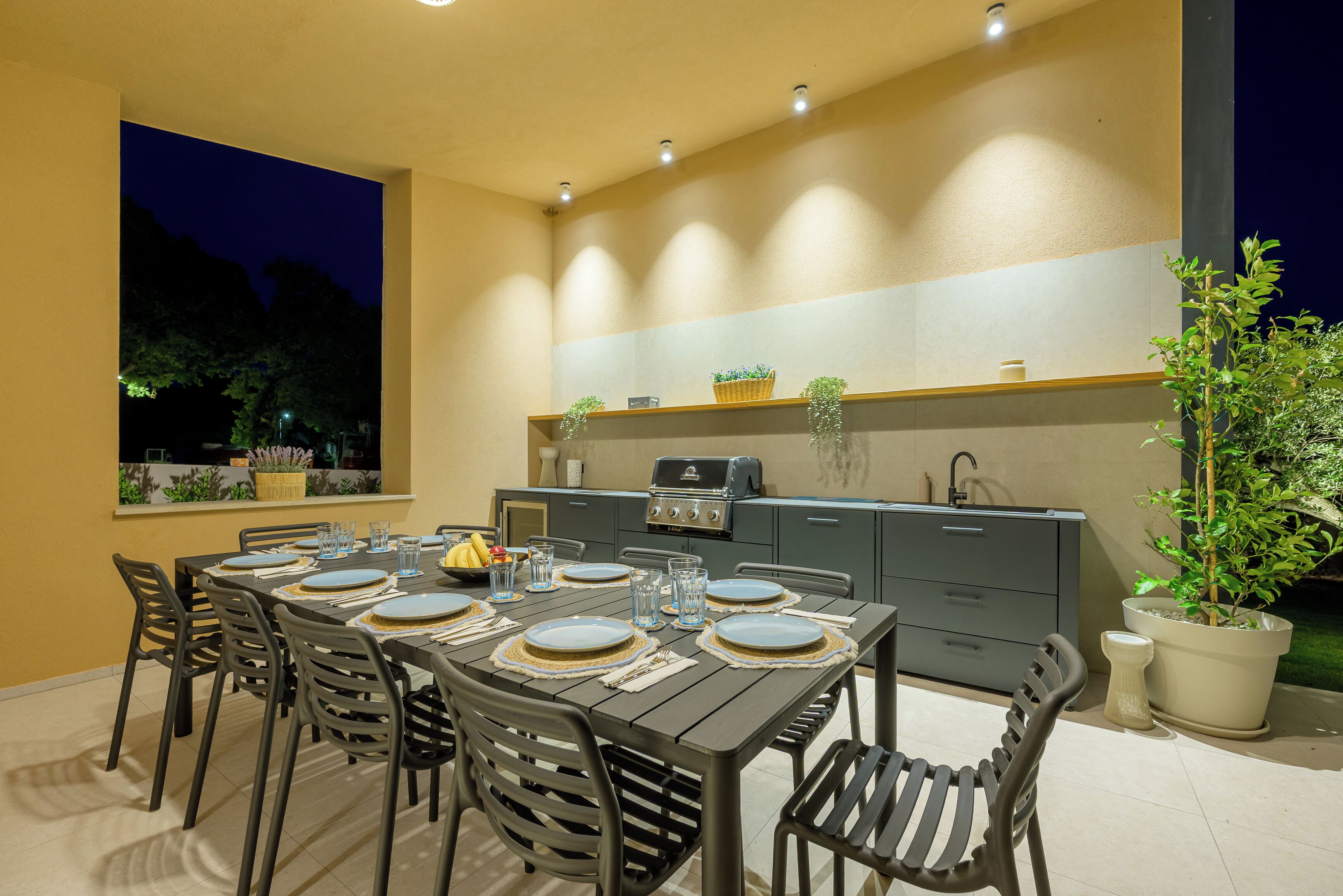 Un área de comedor al aire libre con una mesa preparada para cuatro personas, sillas alrededor y una parrilla al fondo.