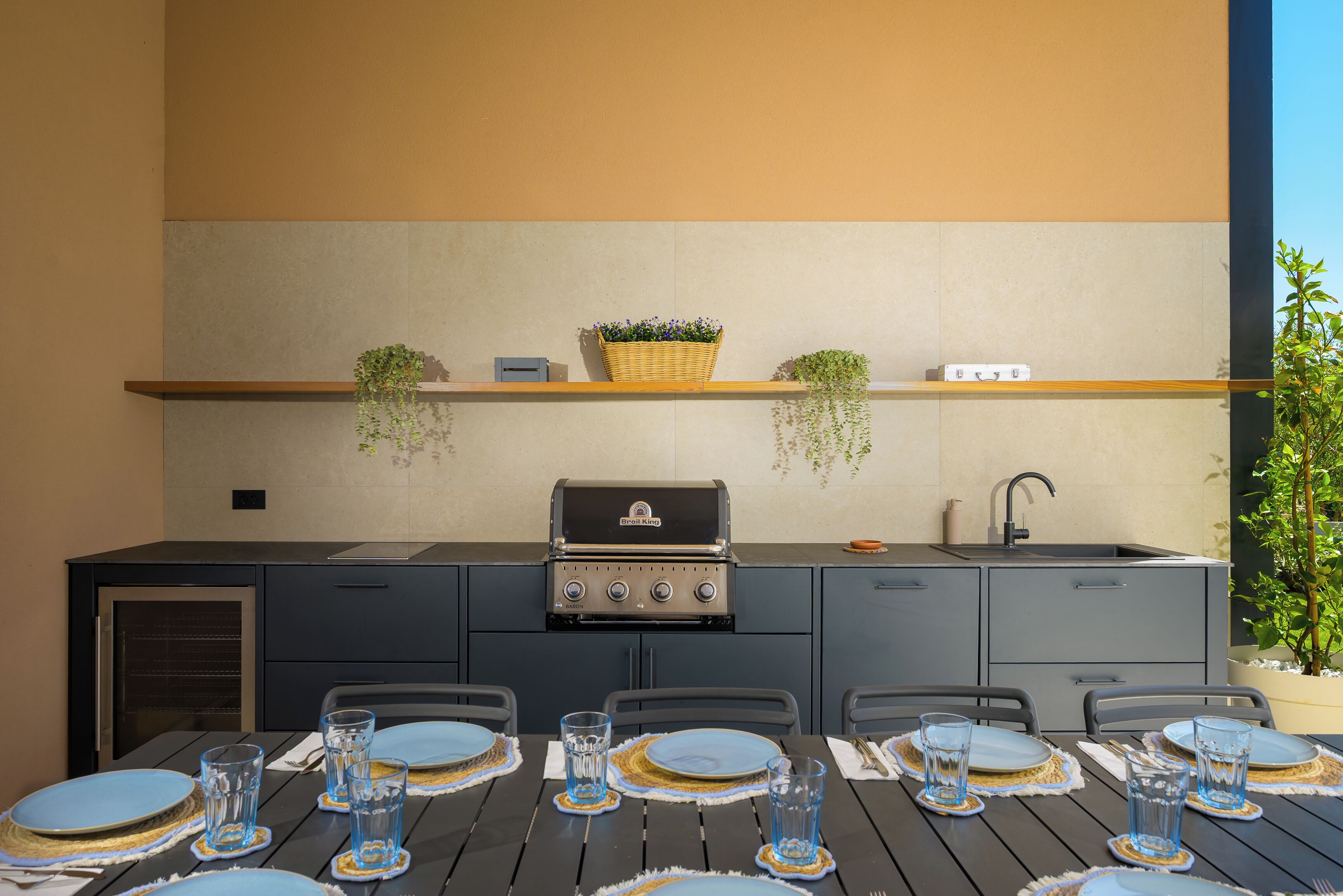 An outdoor kitchen with a grill, shelves with plants, and a dining table set for four.
