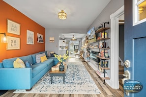 Una sala de estar con un sofá azul, una mesa de centro con libros y un jarrón, un tapete estampado y una estantería con diversos objetos.