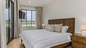 A bedroom with a large bed, a wooden headboard, a TV on the wall, and a window with curtains. - Black Wattle Beach House (Casuarina)