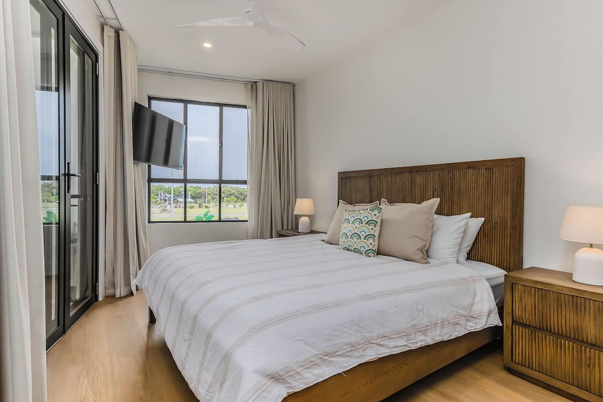 A bedroom with a large bed, a wooden headboard, a TV on the wall, and a window with curtains.
