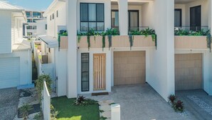 A modern multi-story building with balconies, a central entrance, and a small garden area. - Black Wattle Beach House (Casuarina)