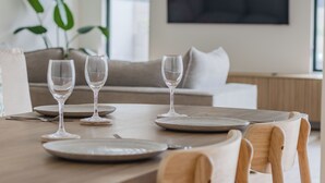 A dining table set with plates and wine glasses, a sofa, and a large window in the background. - Black Wattle Beach House (Casuarina)