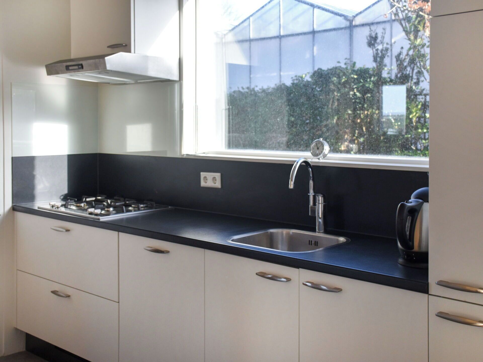 Cocina moderna con estufa a gas, pileta de acero inoxidable y ventana con vista a follaje.