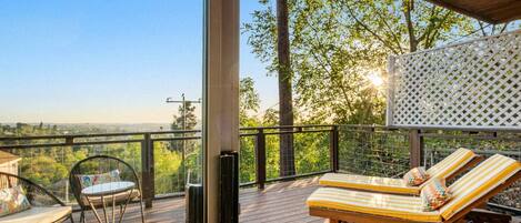 A wooden deck with two lounge chairs and a table, overlooking a scenic landscape.