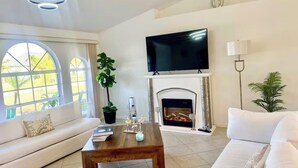 Uma sala de estar com lareira, televisão de tela plana, sofá, mesa de centro e janelas amplas.