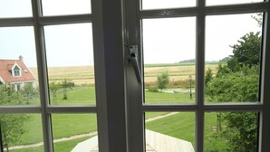 Une fenêtre donnant sur un espace verdoyant, une terrasse en bois et un bâtiment situé au loin.