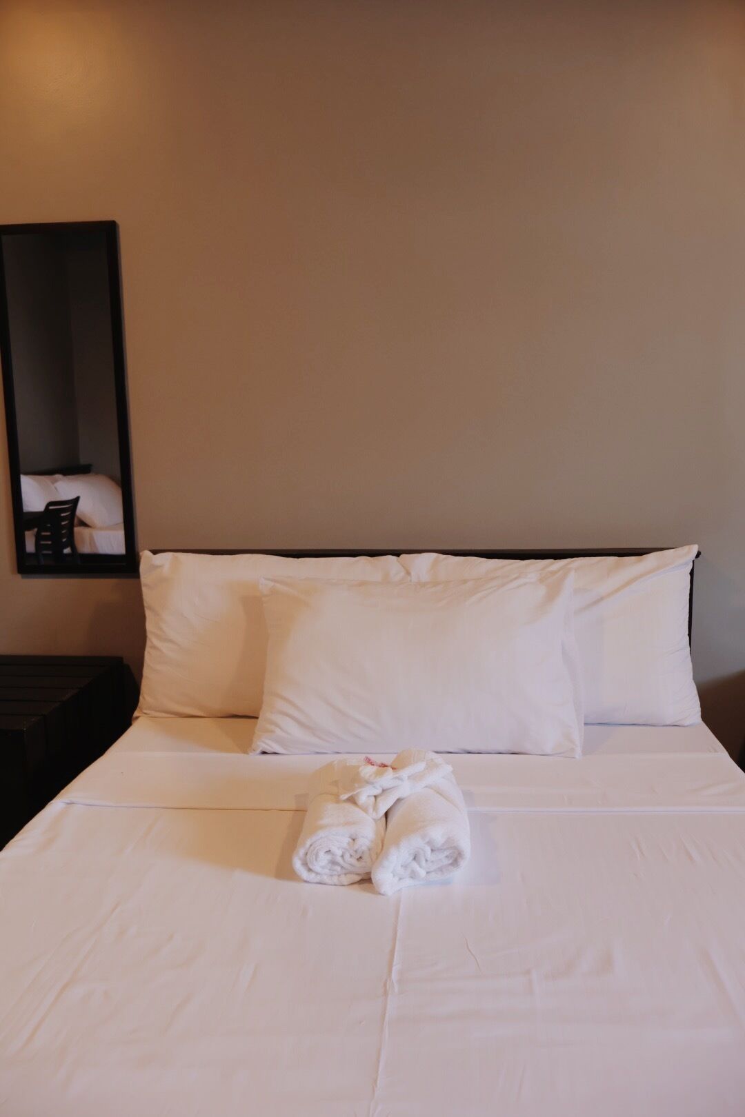 A neatly made bed with white linens and a rolled towel, a dark nightstand, and a mirror on the wall.