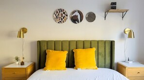 A bedroom with a green upholstered headboard, two yellow pillows, a white bedspread, and a bedside table with a lamp.