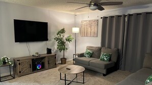 A living room with a flat-screen TV, a ceiling fan, a sofa with green pillows, a coffee table, and a plant. - Comfortable 3BR Home w/ POOL! Near Lackland AFB & Seaworld for BMT Families (San Antonio)