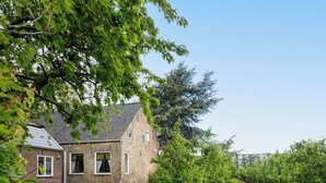 Un bâtiment en briques avec un toit en pente, un petit arbre au premier plan et une rangée d’arbres en arrière-plan.