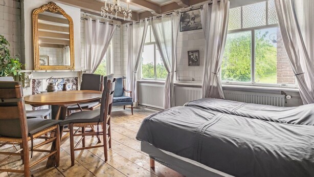 Une pièce comprenant un lit, une table avec des chaises, un lustre et une fenêtre avec des rideaux.
