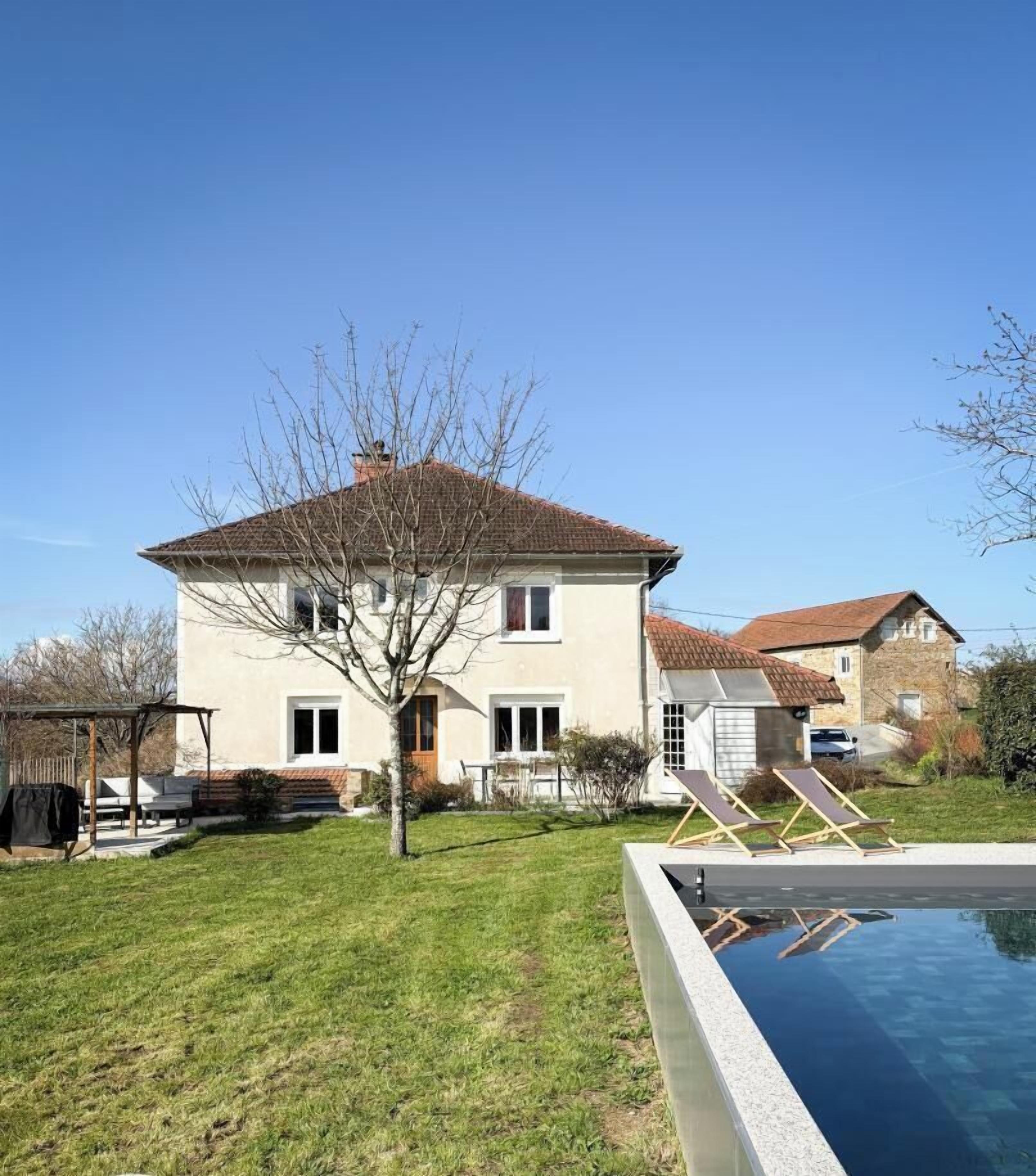 A two-story house with a swimming pool, a patio with chairs, and a small garden.