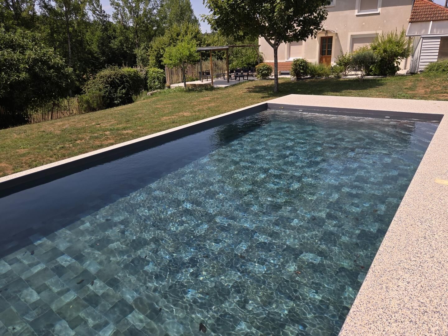 A rectangular swimming pool with clear water, surrounded by a paved area and greenery.