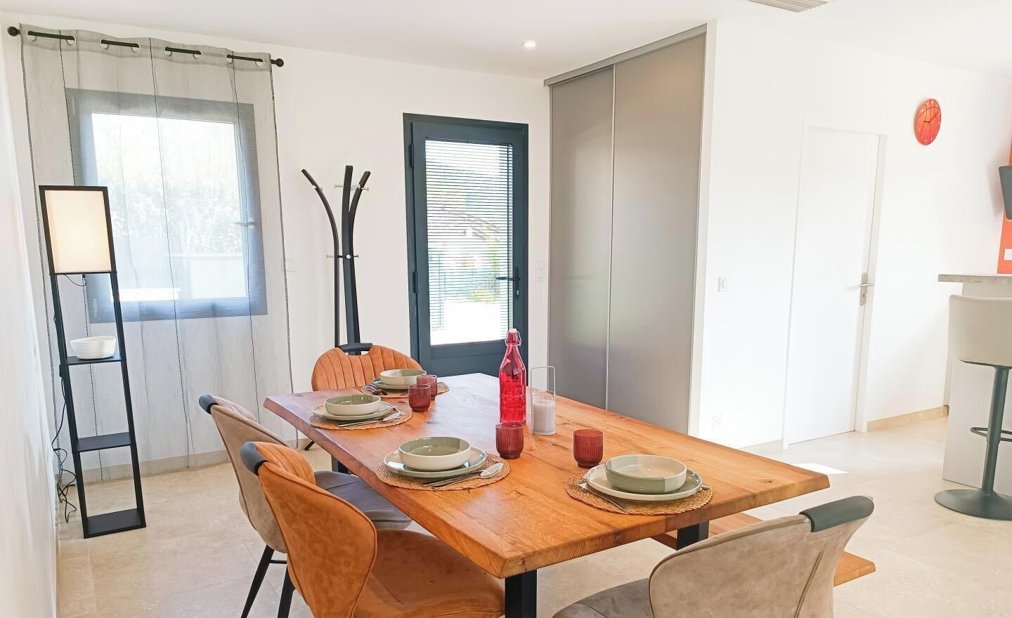 Un coin repas avec une table en bois dressée pour quatre personnes, des chaises, un lampadaire et une porte avec une fenêtre.