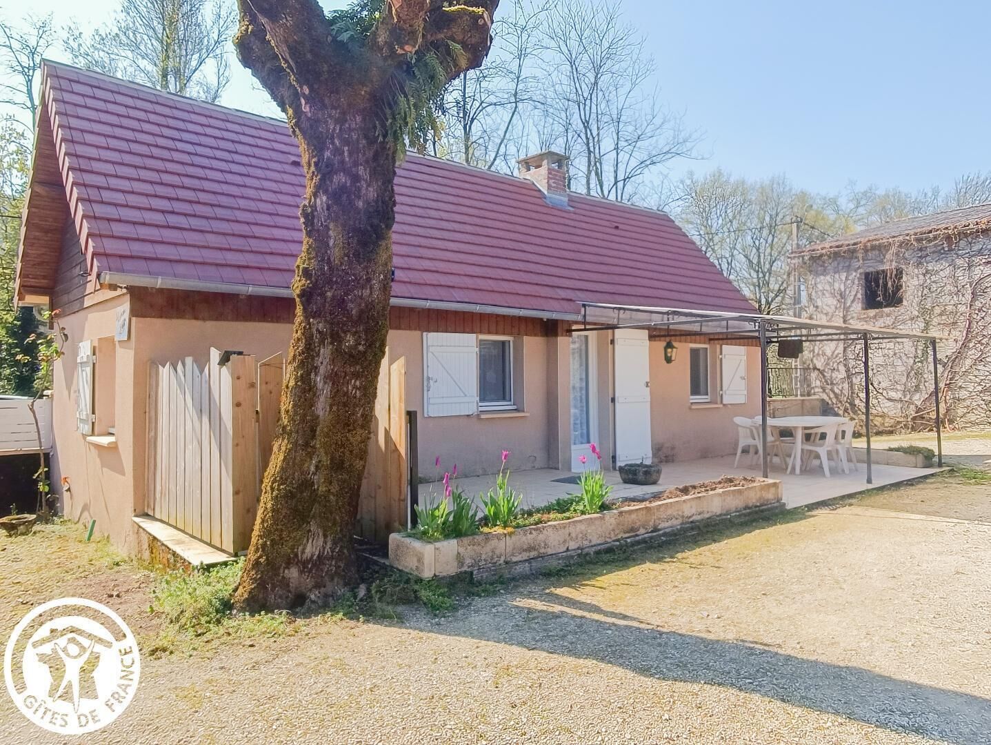 Una casa de una sola planta con techo rojo, cerca de madera y un árbol delante.