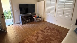 Uma sala de estar com televisão de tela plana, ventilador de teto, tapete estampado, sofá e janela com persianas.