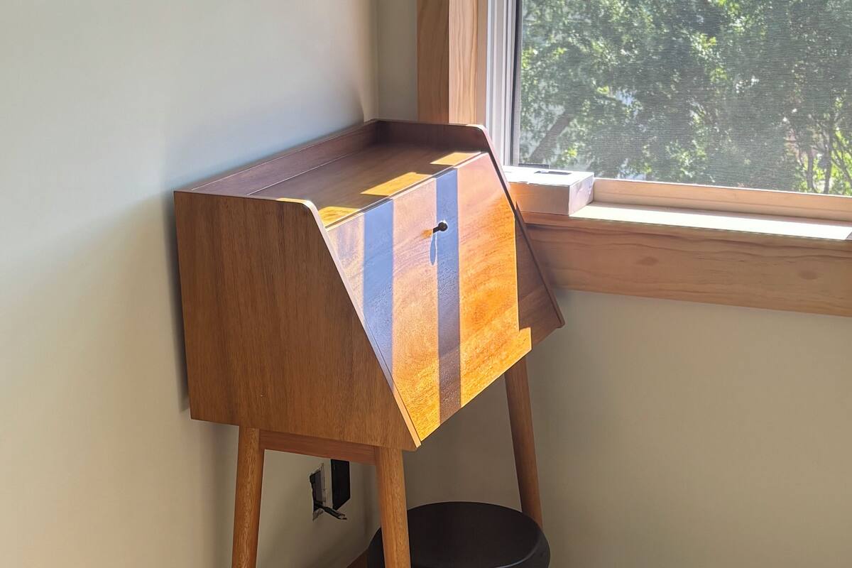 Un bureau d’angle avec un cadre en bois et un tabouret noir, placé près d’une fenêtre donnant sur des arbres.