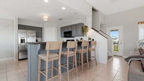 A modern kitchen with a central island, bar stools, and a stainless steel refrigerator. - Serene Coastal Escape With Gulf Views 3 Bedroom Home (Galveston)