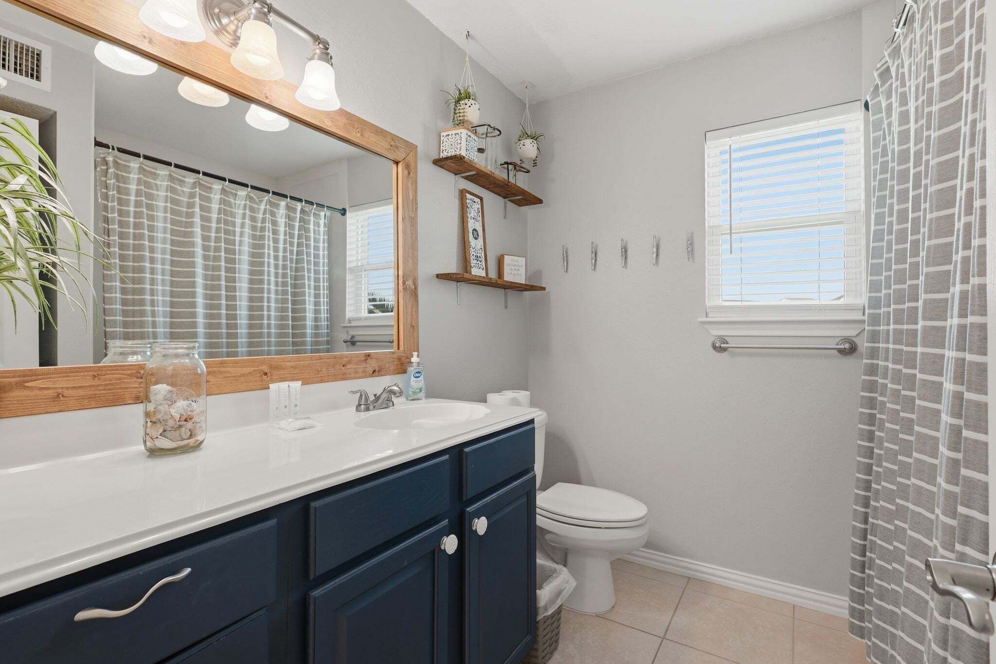 A bathroom with a white sink, dark blue cabinets, a mirror with lights, a toilet, and a window with blinds.