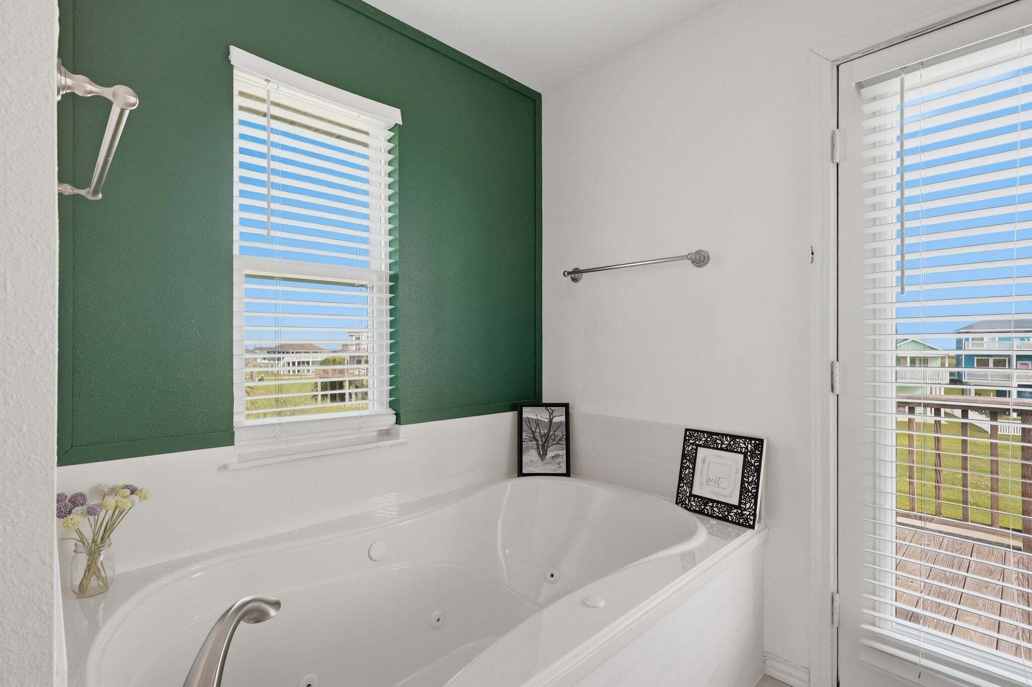 A bathroom with a white bathtub, two windows with blinds, a towel rack, and a framed picture on the wall.