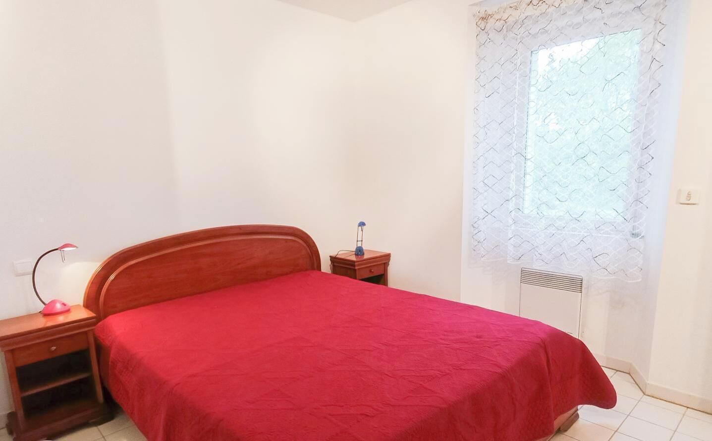 Une chambre à coucher avec un lit en bois, une table de chevet avec une lampe et une fenêtre ornée d’un rideau blanc.
