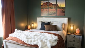 A bedroom with a bed, two bedside tables, a nightstand, a wall-mounted painting, and a window with curtains.