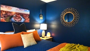Un dormitorio con cama, almohadas, mesita de noche, lámpara, espejo en la pared y un cuadro.