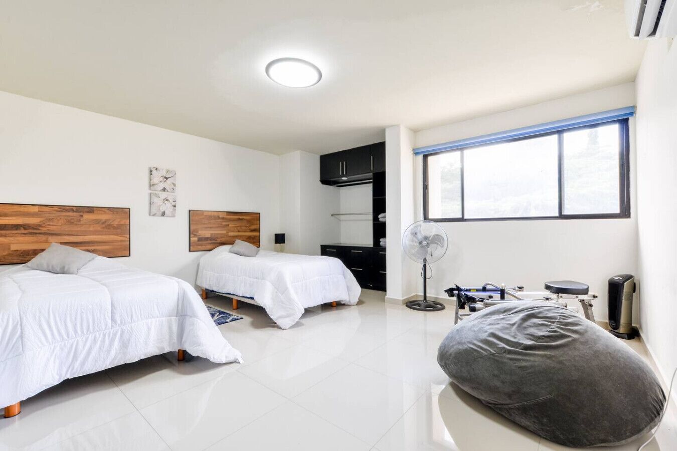 Une chambre d’hôtel moderne avec deux lits, un ventilateur sur pied, un fauteuil massant et une grande fenêtre.