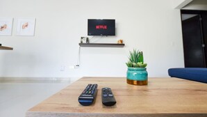 Una sala de estar con una mesa de centro de madera, dos controles remotos, un televisor con soporte en la pared mostrando 'NETFLIX', un estante con una planta y un sofá azul.