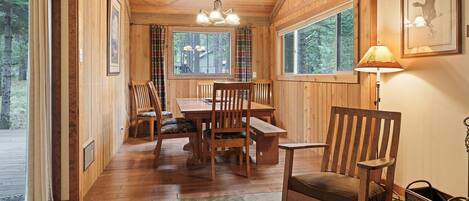 A wooden interior with a dining table, chairs, a rocking chair, a lamp, and a framed picture on the wall.