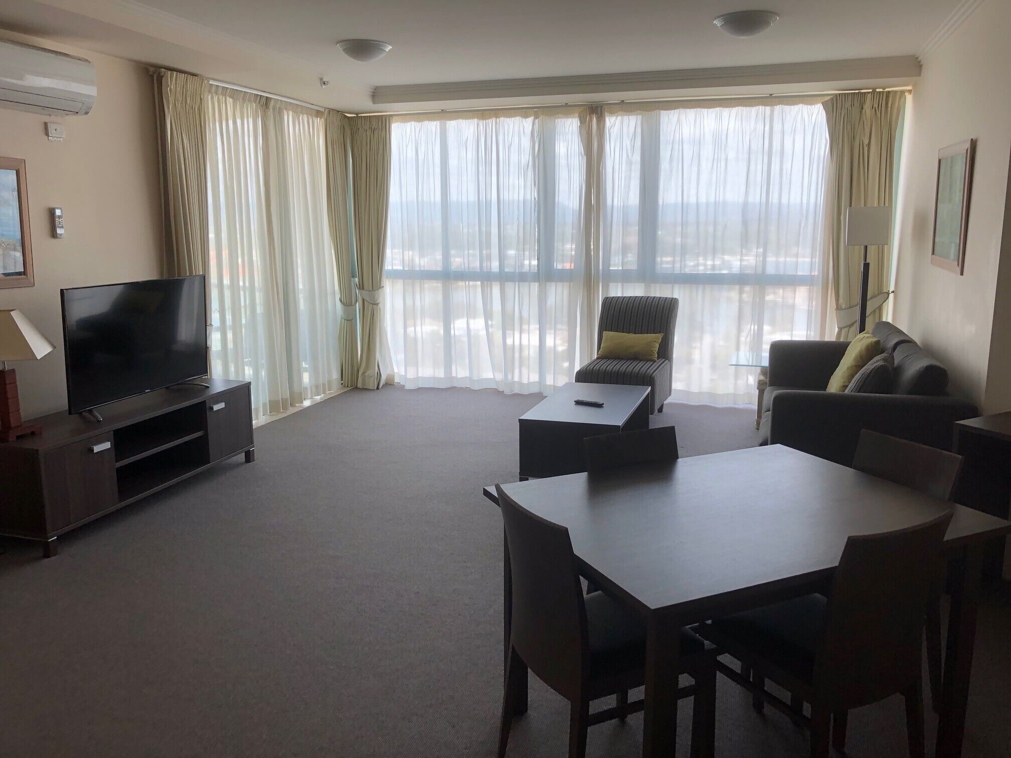 Ein Hotelzimmer mit Flachbildfernseher, Esstisch, Stühlen, einer Couch, einem Balkon mit Vorhängen und einem großen Fenster.