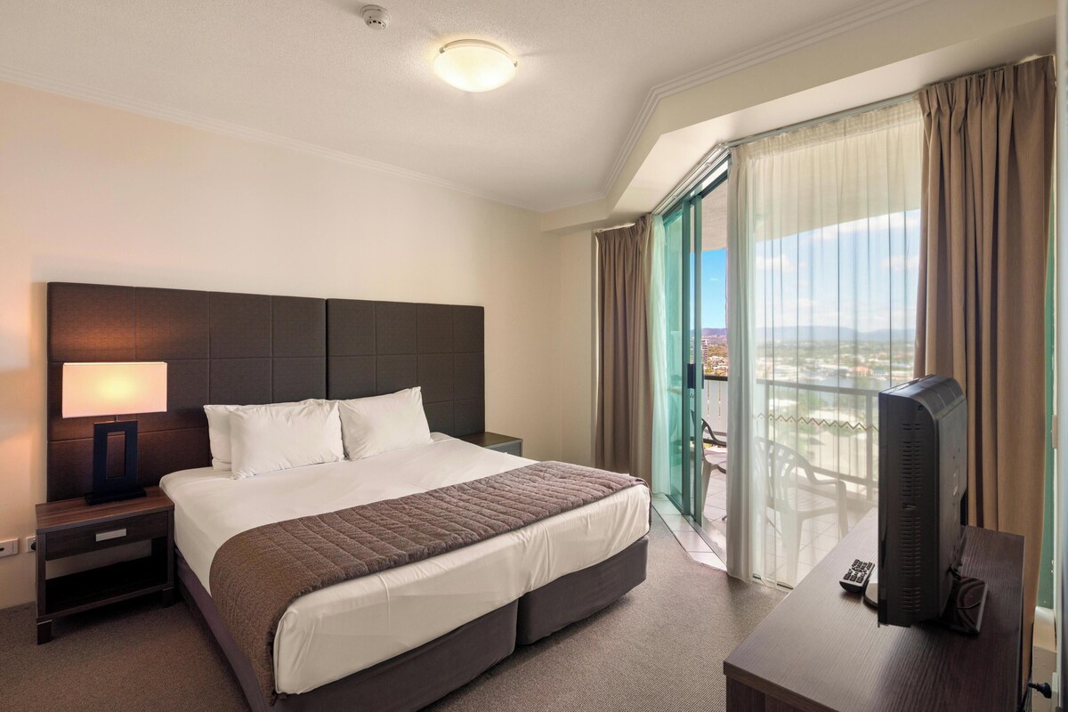 A hotel room with a large bed, a desk with a computer, a nightstand, and a view of the outdoors.