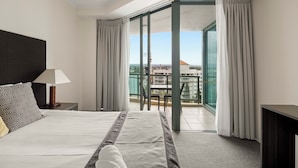 Habitación de hotel con cama, mesita de noche, lámpara, balcón con mesa y sillas, y vista a la ciudad.