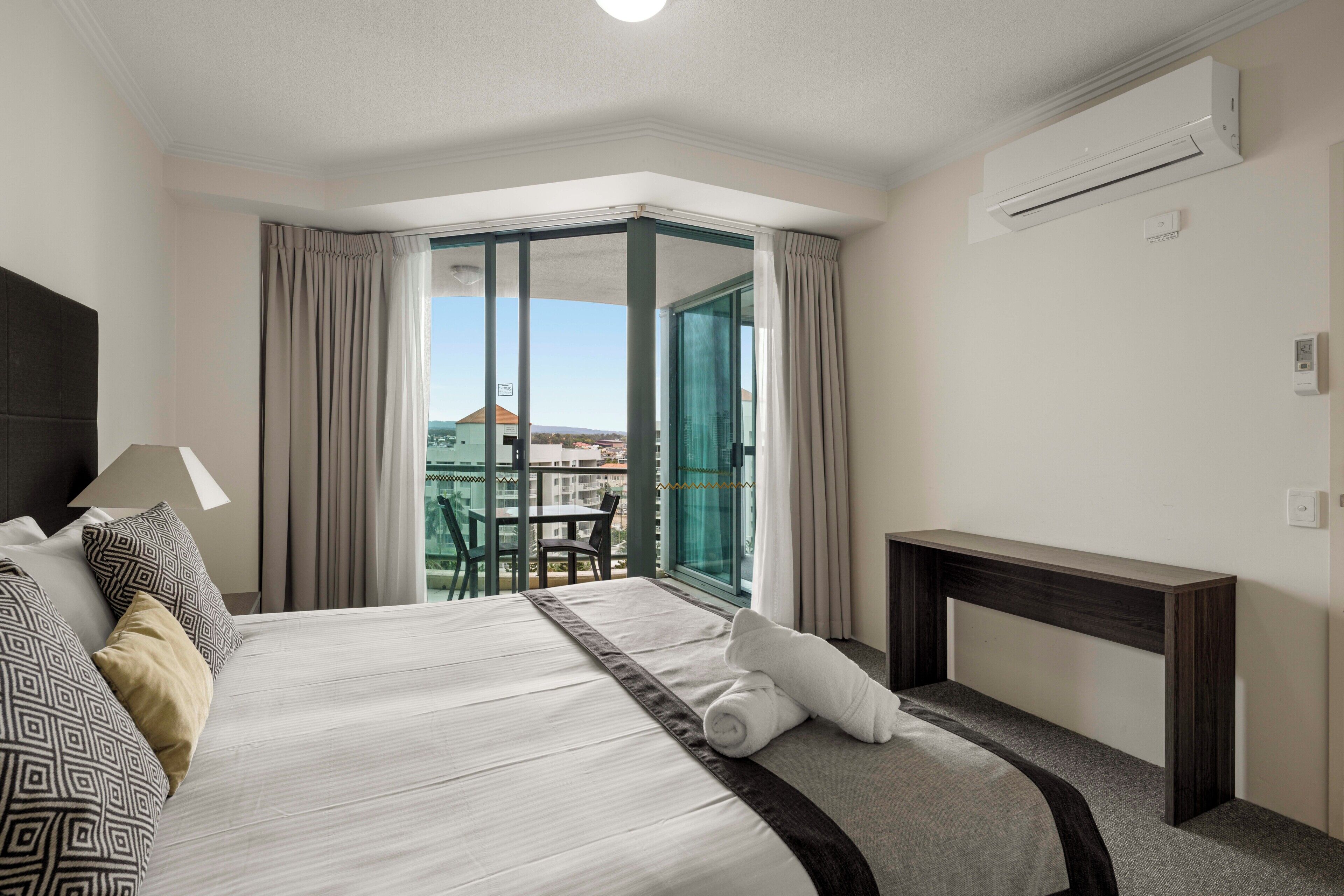 Habitación de hotel con una cama grande, un escritorio, una silla y vistas de la ciudad a través de una puerta de cristal.