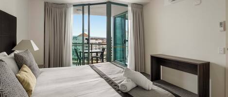 Habitación de hotel con una cama grande, un escritorio, una silla y vistas de la ciudad a través de una puerta de cristal.