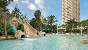 Una piscina con una cascada, rodeada de palmeras y un alto edificio de hotel.