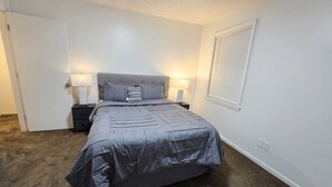 A bedroom with a bed, two bedside tables, a window with blinds, and a door. - Second Home Apartment, Close to Everything (Orangeburg)