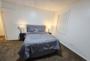 A bedroom with a bed, two bedside tables, a window with blinds, and a door.