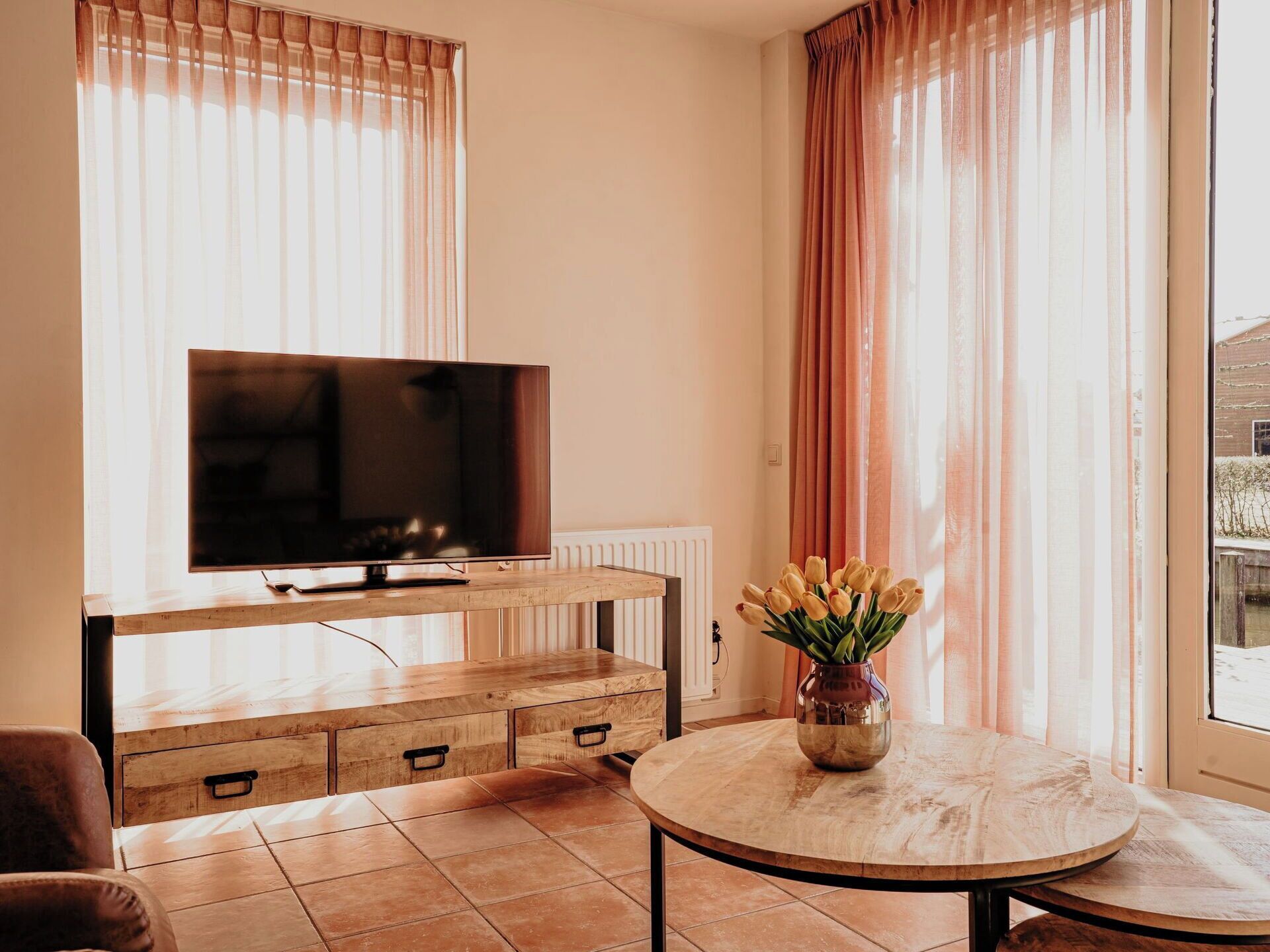 A living room with a TV on a wooden stand, a round table with a vase of flowers, and a window with curtains.