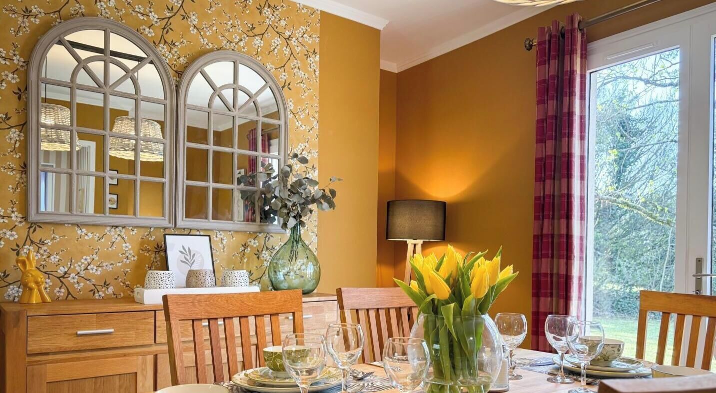 Een eetkamer met een tafel voor vier personen, een vaas met gele bloemen en een raam met uitzicht op groen.