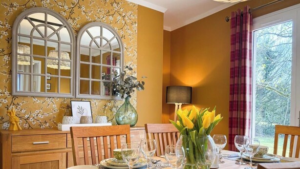 A dining room with a table set for four, a vase of yellow flowers, and a window with a view of greenery. - Le Mûrier - GRAMAT - Gîte (GRAMAT)