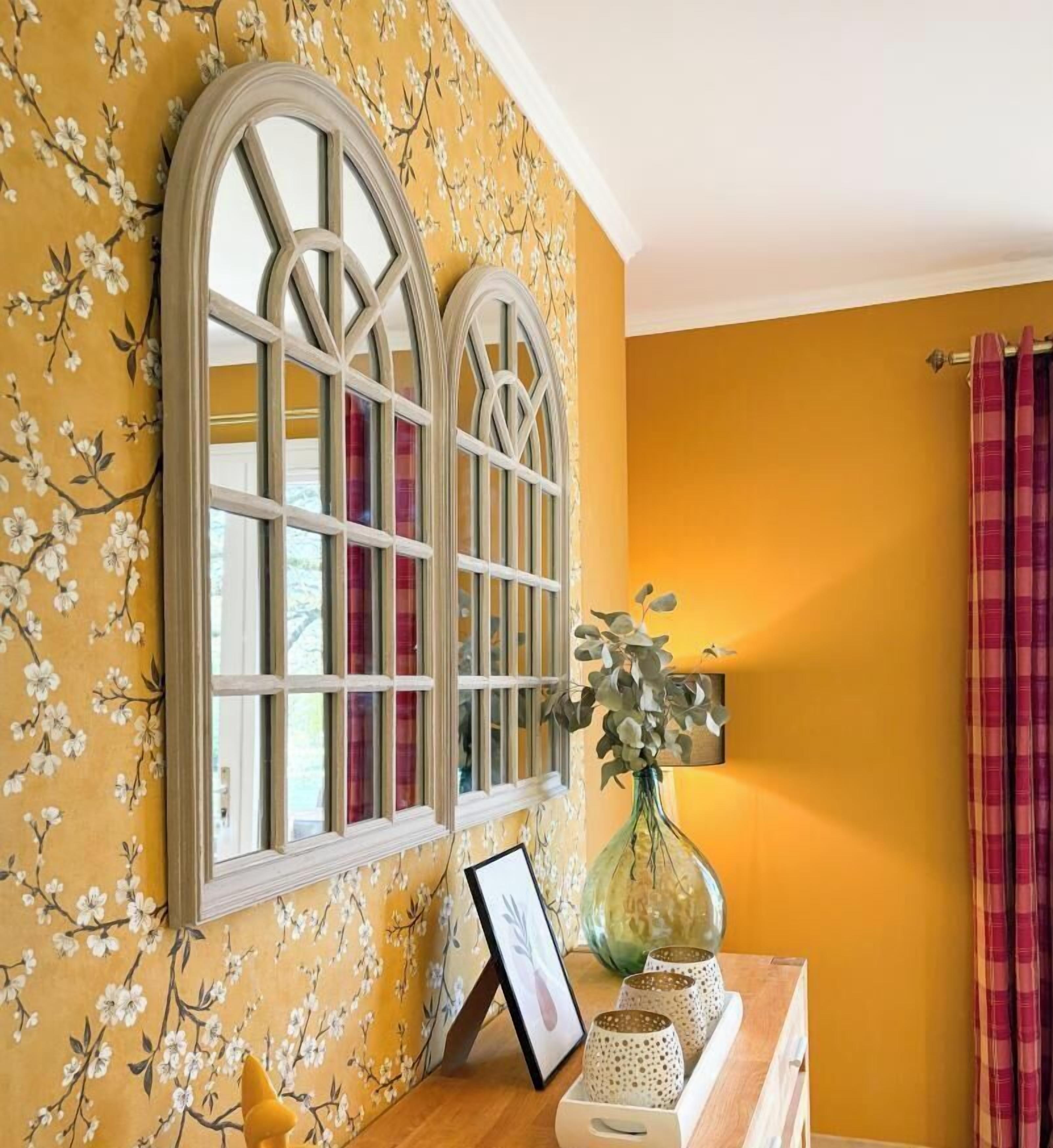Een kamer met bloemenbehang, een houten kastje met een vaas en een fotolijstje, en een raam met gordijnen.