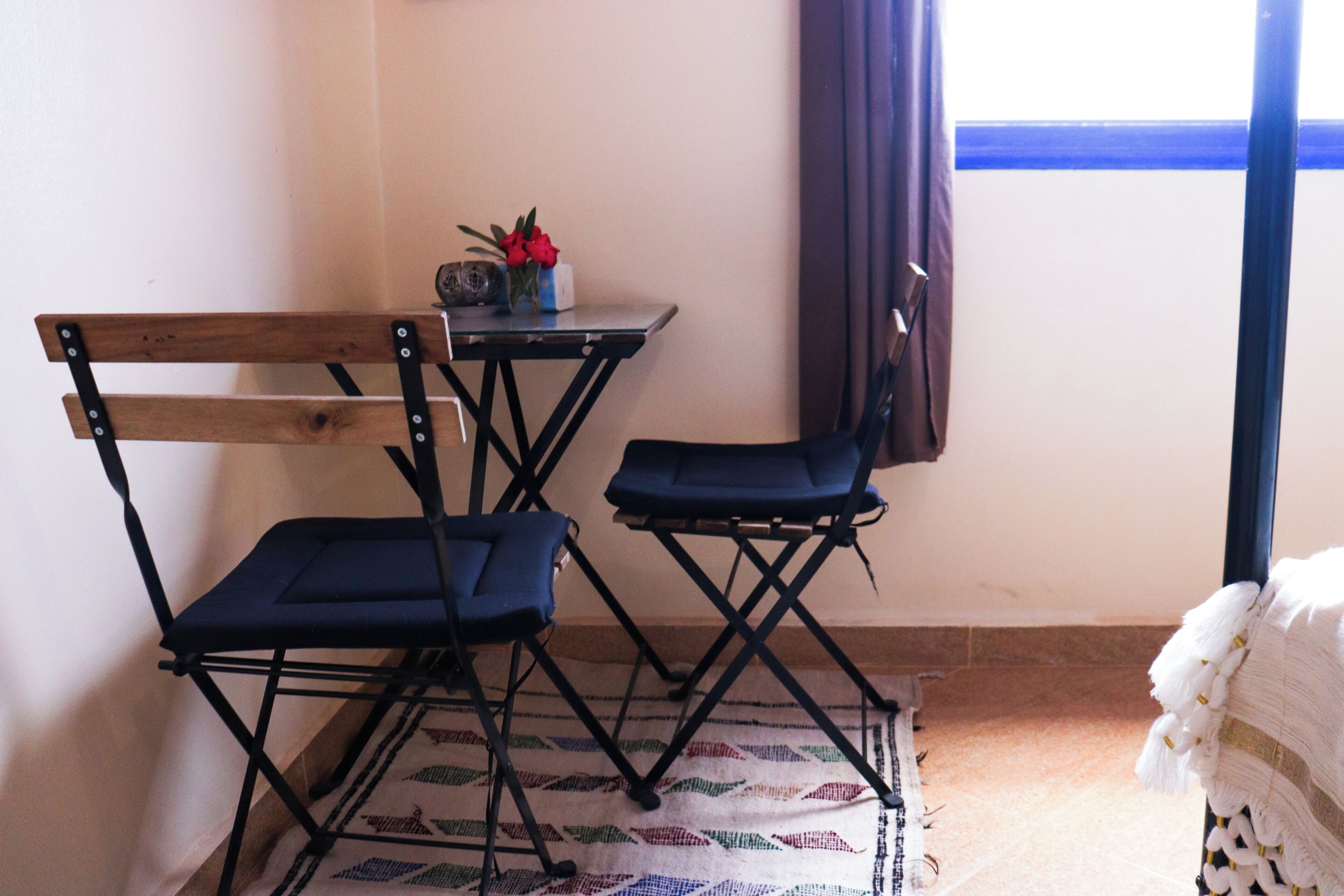 A room with a foldable table and two chairs, a small table with a vase and flowers, a patterned rug, and a window with curtains.