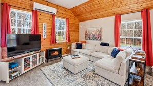 Un intérieur de chalet chaleureux avec un téléviseur, une cheminée, un canapé et de grandes fenêtres.