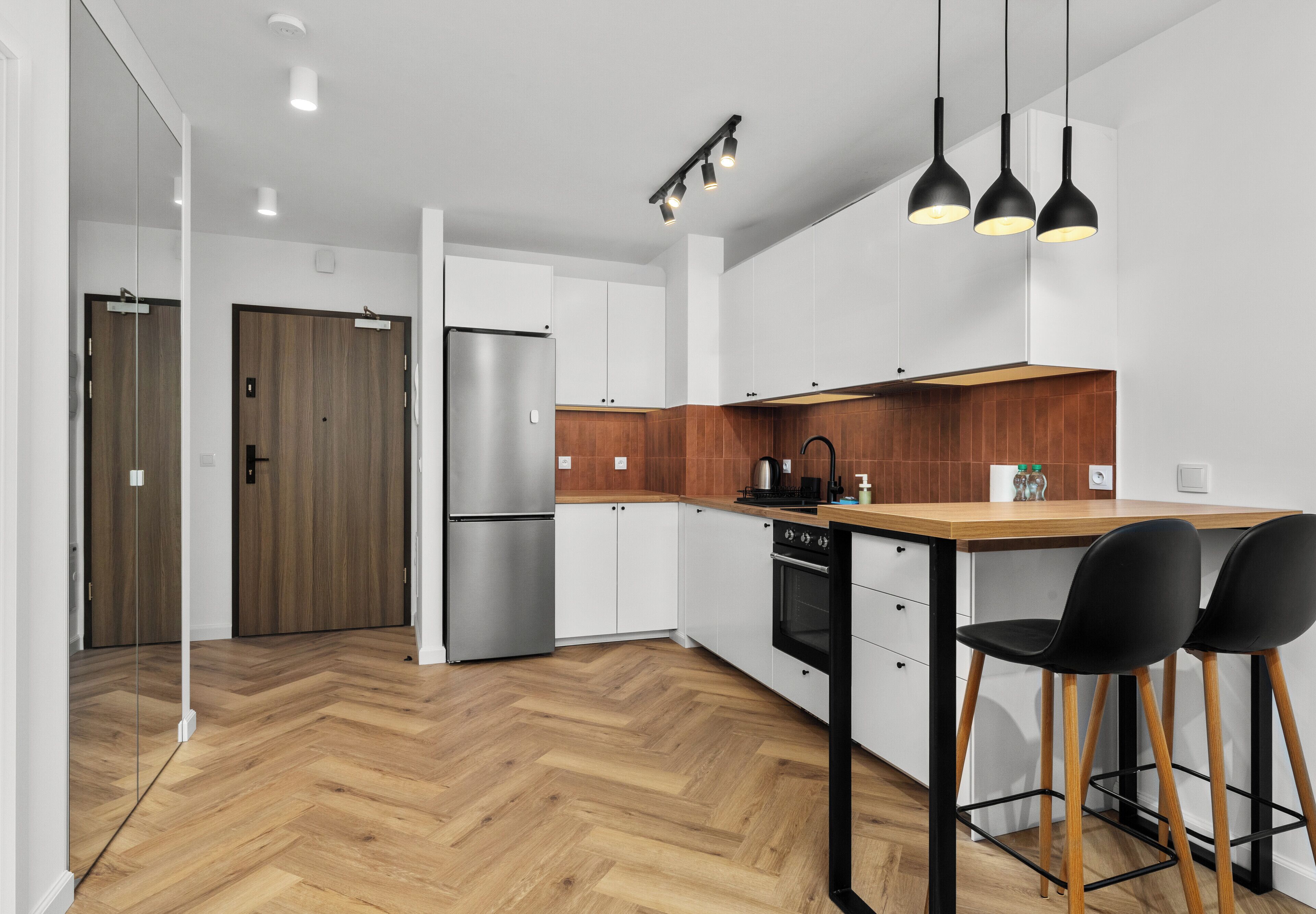 A modern kitchen with a wooden herringbone floor, white cabinets, a stainless steel refrigerator, a wooden countertop, and black bar stools.