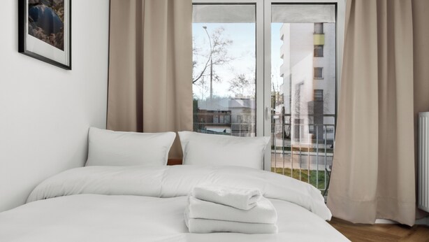 Un lit bien fait, avec du linge de lit blanc et une serviette pliée, une fenêtre avec des rideaux et un tableau encadré accroché au mur.